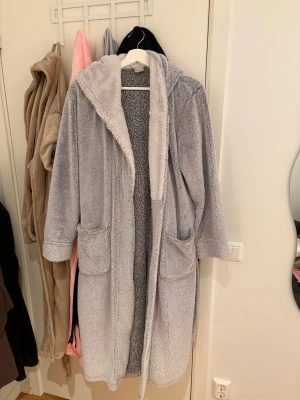 Ljusgrå fluffig morgonrock med huva - Mjuk och fluffig ljusgrå morgonrock med stor huva och två rejäla fickor framtill. Den har ett matchande bälte i midjan och är tillverkad i gosig fleece som känns riktigt skön mot huden. Perfekt för att mysa runt hemma under kyliga dagar.