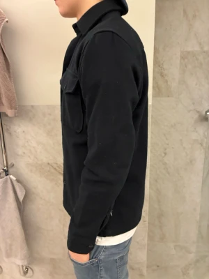 Svart overshirt med fickor - Säljer en svart overshirt med klassisk krage och två stora bröstfickor med knappar. Jackan har en rak och normal passform, är långärmad med knappar. Väldigt stilren och funkar till mycket. Hör av dig vid frågor !