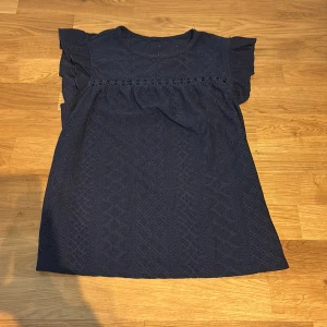 Mörkblå blus med spets och volang - Supersöt mörkblå blus med spetsdetaljer och volangärmar från shein. Blusen har rund halsringning och ett fint broderat band med blommor över bröstet. Materialet är lätt och luftigt med broderat mönster över hela toppen.