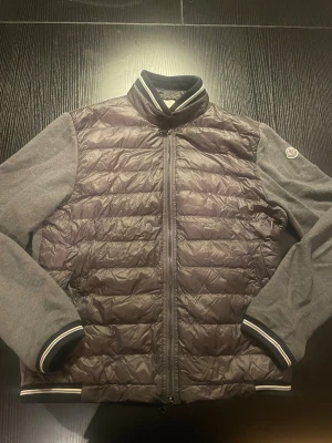 Grå quiltad cardigan jacka Moncler - Snygg grå cardigan jacka från Moncler med quiltad pufferfront och mjuka stickade ärmar. Jackan har en defekt som synd på ( BILD 6). 