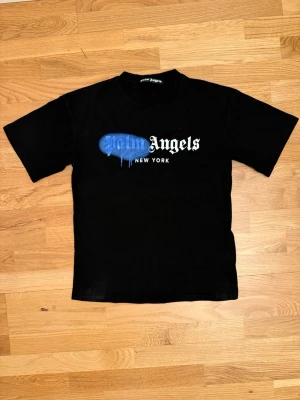 Palm Angels svart t-shirt  - Svart t-shirt från Palm Angels med tryck på bröstet där det står 'Palm Angels New York' i vitt och blått. Den har en klassisk passform, rund hals och korta ärmar. Materialet är mjuk bomull och trycket har en cool drip-effekt.
