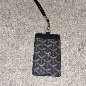 Svart  korthållare med mönster - Säljer en svart korthållare från  med det klassiska grafiska mönstret i vitt och grått. Korthållaren har en praktisk rem och transparent fack på baksidan för ID eller kort. Perfekt accessoar för dig som vill ha koll på korten med stil.