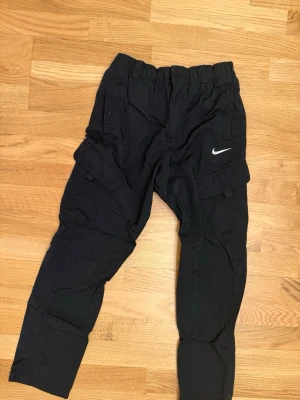 Svarta cargopants från Nike, storlek L - Svarta cargopants från Nike med elastisk midja och flera fickor på benen. Snyggt Nike-logga broderad på ena benet. Byxorna är gjorda i ett lätt och slitstarkt syntetmaterial, perfekta för en chill och sportig stil.