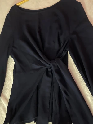 Svart långärmad blus med knyt - Säljer en svart långärmad blus med snygg knytning i midjan. Blusen har en lös passform och är tillverkad i ett tunt, mjukt tyg som faller fint. Perfekt för dig som gillar stilrena plagg med en twist.