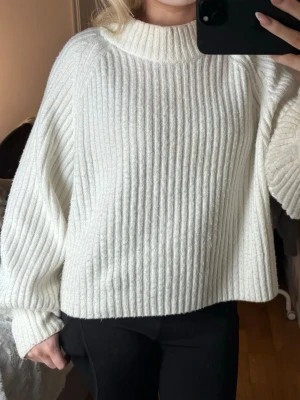 Vit ribbad stickad tröja - Säljer en vit stickad tröja med ribbat mönster och hög hals. Tröjan har en loose passform och långa ärmar som ger en avslappnad vibe. Perfekt för kyliga dagar och enkel att styla med jeans eller kjol.