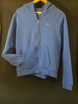Blå Lacoste Zip - En blå Lacoste zip är en stilren och bekväm tröja med hel dragkedja. Den har en mjuk kvalitet, sportig design och den ikoniska krokodillogga. Inga defekter.Perfekt för en avslappnad men välklädd vardagsstil