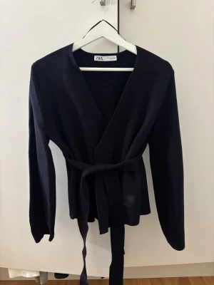 Mörkblå omlottkofta från Zara - Säljer en mörkblå omlottkofta från Zara i storlek S. Koftan har långa ärmar och ett matchande bälte i midjan för att knyta omlott. Materialet är mjukt och tjockt. Perfekt för lager på lager under höst och vinter.
