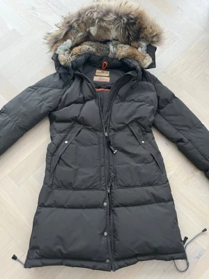 Parajumpers lång svart dunjacka S - Säljer en lång svart dunjacka från Parajumpers med stor huva och fluffig pälsdetalj. Jackan har dragkedja, knappar, två stora fickor och snygg patch på ärmen. Perfekt för kalla vinterdagar och riktigt varm.