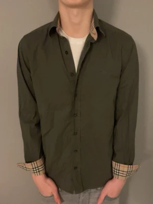 Burberry skjorta - Mörkgrön skjorta från Burberry med klassiska beige-rutiga detaljer på insidan av krage och manschetter. Skjortan har knappar framtill och lång ärm. Perfekt för dig som vill ha en clean look med en twist av mönster. Modellen är ca 180cm och väger 67kg.