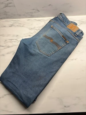Blå jeans från Nudie Jeans - Märke: Nudie Jeans.                                           Modell: Lost orange Passform: Rak / slim (utifrån bilderna: klassisk rak–slim passform).                Färg: Blå denim Skick: Bra begagnat skick     Snygga Nudie Jeans i klassisk blå tvätt. Tidlös modell som funkar till både vardag och mer uppklätt. Jeansen är i fint skick med naturligt slitage som ger en snygg denimlook. Inga hål eller fläckar. Tillverkade i kvalitetsdenim som håller länge – 