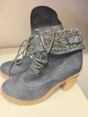 Blå stövletter med snörning och klack - Snygga blå stövletter med grov klack och snörning framtill. Ovandelen är i mockaliknande material och har en gråmelerad, ullig kant med spänndetalj. Sulan är beige och rejäl, perfekt för höst och vinter. Skorna har rund tå och cool outdoor-vibe.