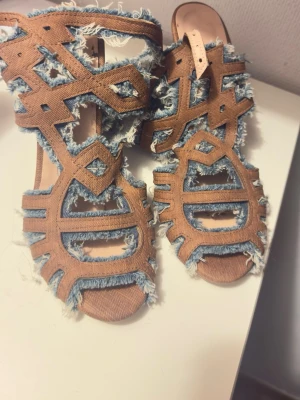 Bruna sandaler med denim och klack - Unika sandaler från Roc med chunky klack och öppen tå. Skorna har ett coolt mönster i brunt tyg med slitna denimdetaljer och fransiga kanter. Perfekta för dig som vill sticka ut med en edgy och trendig look.