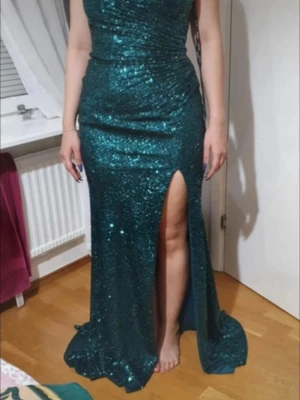 Grön glittrig långklänning med slits - Säljer en smaragdgrön långklänning med paljetter och smala axelband. Klänningen har en djup slits framtill, omlottkänsla och snörning i ryggen. Perfekt för bal eller fest. Materialet är glittrigt och skimrande, vilket ger en lyxig känsla.