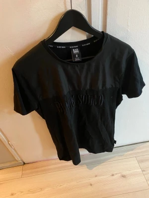Svart t-shirt från Black Squad - Säljer en svart t-shirt från Black Squad i regular fit. T-shirten har korta ärmar och ett diskret broderat Black Squad Paris-tryck på bröstet. Tillverkad i mjuk bomull som känns skön mot huden. Enkel och clean design som passar till allt.