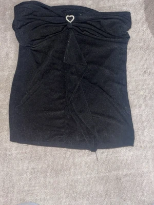 Svart bandeau topp med hjärtspänne - Säljer en svart bandeau topp i mjukt tyg med en glittrig hjärtformad detalj framtill. Toppen har en snygg drapering och en liten slits framtill som ger extra edge. Perfekt för dig som vill ha en stilren men ändå unik look.