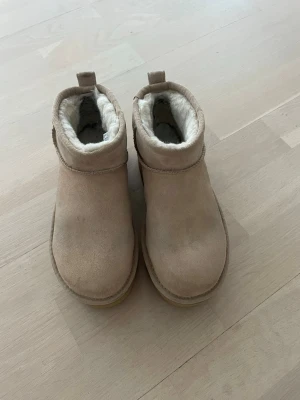 Beige UGG boots med plattform - Säljer ett par beige UGG boots med plattformsula och rund tå. Skorna är fodrade med mjuk, vit päls på insidan och har en ovandel i mocka. Klassisk UGG-logga bak på hälen och dragflik för enkel påtagning. Perfekta för kalla dagar.