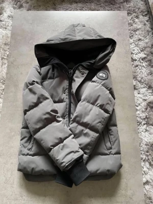 Canada goose macmillian - Säljer en grå pufferjacka från Canada goose med stor huva och svart innerfoder. Jackan har dragkedja framtill, två sidofickor och en patch på ärmen. Perfekt för kalla dagar och riktigt skön att bära.