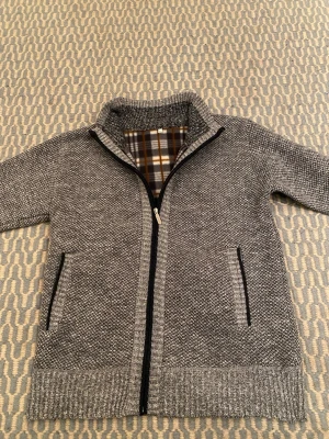 Grå stickad kofta med dragkedja - Mysig grå stickad kofta med hel dragkedja och två fickor fram. Insidan har rutigt foder i brunt, vitt och grått som ger extra värme. Ribbstickade muddar vid ärmslut och nederkant. Perfekt för kyliga dagar och enkel att matcha. Står som M men sitter som S