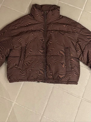 Brun mönstrad pufferjacka - Säljer en brun pufferjacka med unikt vågigt mönster i olika bruna toner. Jackan har hög krage, dragkedja framtill och två stora fickor. Perfekt för dig som vill sticka ut i vinter och hålla dig varm med stil.
