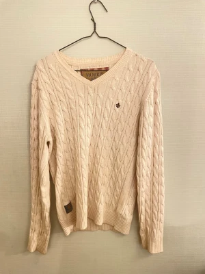 Kabelstickad V-neck  Från Morris - Kabelstickad beige tröja från Morris med v-ringning och diskret logga broderad på bröstet. Tillverkad i 100% bomull och har ribbade muddar vid ärmslut och nederkant. Perfekt för lager på lager under höst och vår. 