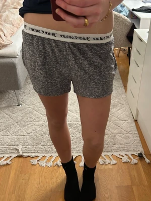 Juicy Couture gråa mjukisshorts  - Mörkgrå melerade shorts från Juicy Couture med bred resår i midjan där loggan står tryckt runtom. På baksidan finns texten 'Juicy' i glittriga strassdetaljer. Mjukt och stretchigt material, perfekta för chill dagar.