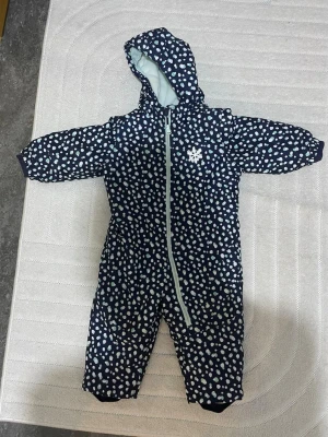 Mörkblå täckoverall med prickar - Mysig mörkblå täckoverall med vitt och ljusblått prickmönster. Overallen har huva, mjukt ljusblått foder och dragkedja framtill. Perfekt för kalla dagar och håller dig varm och bekväm. Snygg detalj med snöflinga på bröstet.