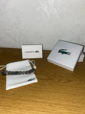 Lacoste Armband - Bilderna visar ett armband från Lacoste i silverfärg med tillhörande ask och påse, men ingen väst syns på bilderna. Inga repor!!!