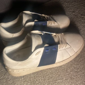 Valentino Garavani vita sneakers - Säljer ett par vita Valentino Garavani sneakers med bred marinblå rem över vristen och matchande blå detaljer vid snörningen. Skorna har klassisk rund tå, platt sula och små nitar längs hälen. Tillverkade i skinn och har snörning framtill.