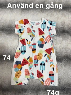 Färgglad byxdress med glassmönster - Söt byxdress i vitt med färgglatt mönster av glassar, vattenmeloner och ananas. Kortärmad och med tryckknappar i grenen för enkel påklädning. Perfekt för varma dagar och riktigt lekfull stil.🌸 Använd en gång. Storlek 74 