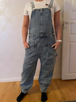 Baggy denim byxdress med stora fickor - Säljer en baggy blå jeans, byxdress med stora fickor framtill och justerbara axelband. Byxorna har en loose passform med spännen nertill. 