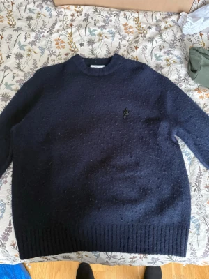 Axel arigato - Säljer en mörkblå stickad tröja från  Axel arigato  med rund hals och diskret logga broderad på bröstet. Tröjan har långa ärmar och ribbade muddar vid nederkant och ärmslut. Perfekt för kyliga dagar och enkel att matcha med jeans.