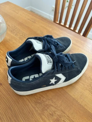 Converse CONS blå sneakers  - Säljer ett par Converse CONS sneakers i mörkblå mocka med vita detaljer och snörning. Skorna har vit sula, klassisk stjärnlogga på sidan och CONVERSE-märke på plösen. Insidan är blå med stor logga. Perfekta för dig som gillar streetwear och retrostil. De är dessutom i väldigt gott skick och med två olika färg på snörningen. Vid frågor är det bara att skriva! Kom med bud!