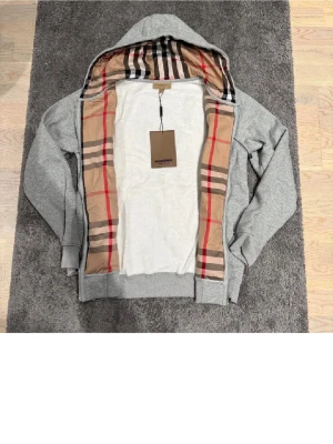 Grå Burberry hoodie med dragkedja - Säljer en grå hoodie från Burberry med klassiskt rutigt foder i huvan och insidan. Tröjan har dragkedja, snörning i huvan och diskret broderad logga på bröstet. Tillverkad i mjuk bomull och har ribbade muddar vid ärmar och nederkant.