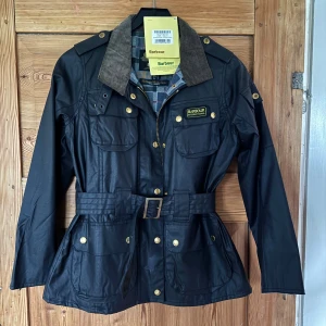 Svart Barbour jacka - Svart Barbour International jacka med bälte i midjan, guldfärgade knappar och dragkedja. Klassisk krage i brun manchester och rutigt foder i grått, blått och orange. Fyra fickor framtill och Barbour-logga på bröstet. Perfekt för dig som gillar stilren och funktionell design.