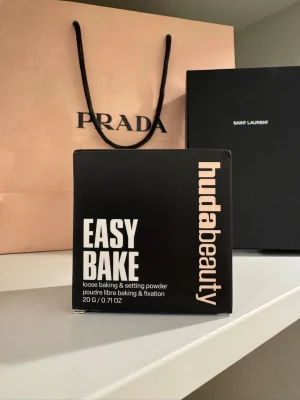Huda Beauty Easy Bake Banana Bread - Loose baking & setting powder i nyansen Banana Bread från Huda Beauty. Kommer i en svart förpackning med beige etikett. Pudret är lätt och silkeslent, perfekt för att fixera sminket och ge en matt finish. Innehåller 20 g och appliceras med borste eller beautyblender.