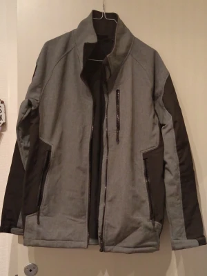 Grå vindjacka från Bluewear M - Snygg grå vindjacka från Bluewear med svarta paneler på sidor och ärmar. Jackan har hög krage, tre dragkedjefickor och är tillverkad i 100% polyester. Perfekt för dig som gillar stilren och funktionell design.