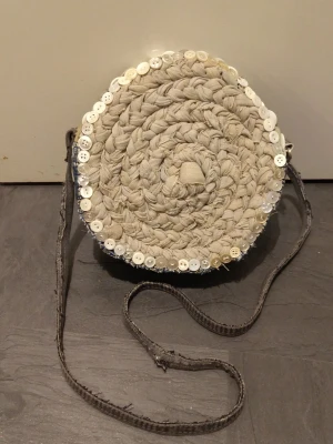 Rund axelväska i denim och beige tyg - Unik rund axelväska med framsida i flätat beige tyg och kantad med vita knappar. Baksidan och sidorna är gjorda av blå denim med synliga sömmar. Väskan har en lång, smal axelrem i mörkgrått tyg. Perfekt för dig som gillar kreativ och personlig stil. Återbruk av gamla jeans