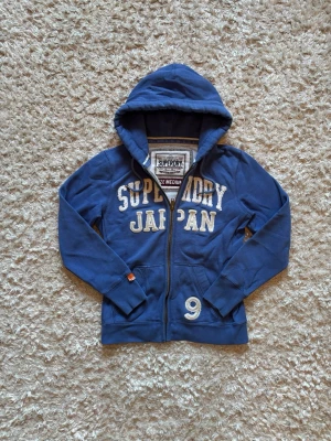 Blå zip hoodie från superdry - Snygg blå hoodie från superdry, den är använd men i bra skick. Storlek M men sitter absolut mindre så rekommenderar att jämföra med måtten nedan!                                                       Längd: 60 Ärm: 61 Bröstvidd: 50