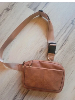 Brun axelväska i skinn med dragkedja - Säljer en brun axelväska i skinn med justerbart band och praktiskt spänne. Väskan har ett stort fack med dragkedja och en extra ficka framtill. Perfekt storlek för mobil, plånbok och nycklar. Enkel och stilren design som funkar till många outfits.