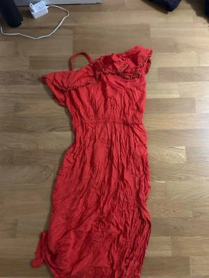 Röd långklänning med volang  - Säljer en röd långklänning från Lindex med volangdetaljer vid axlarna och en axelbandslös design på ena sidan. Klänningen har en smal passform och är tillverkad i mjuk bomull. Perfekt för dig som gillar färgstarka och trendiga plagg.