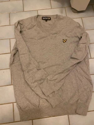 En grå tröja från Lyle & Scott - Säljer en grå tröja från Lyle & Scott i storlek M. Tröjan har v-ringad hals och ett broderat gult fågelmärke på bröstet. Mjukt material och ribbade muddar vid ärmslut och nederkant. Perfekt för dig som gillar stilrena och klassiska plagg.