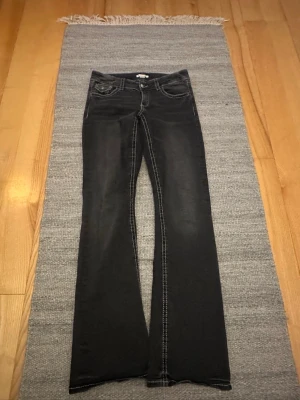 Svarta bootcut jeans från Gina Tricot strl 170 - Svarta jeans med bootcut-snitt från Gina Tricot. Snygga kontrastsömmar i vitt och klassiska fem fickor med lock och knapp på bakfickorna. Jeansen har låg midja och är gjorda i stretchig denim för skön passform.