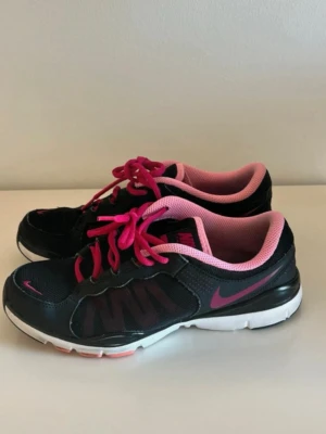 Nike sneakers svart och rosa strl 37.5 - Säljer ett par snygga Nike sneakers i svart med rosa detaljer och rosa snörning. Skorna har mesh- och syntetmaterial, vit sula och rund tå. Perfekt för dig som gillar sportig stil och vill ha bekväma skor med cool färgkombo.