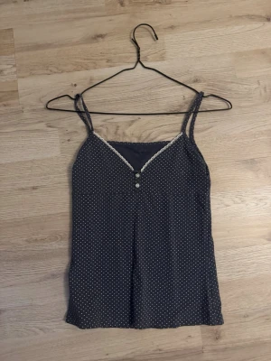 Mörkblå prickig linnetopp med spets brandy Melville - Säljer en mörkblå linnetopp med vita prickar och tunna axelband. Toppen har v-ringad hals med vit spetskant och två dekorativa knappar framtill. Perfekt för varma dagar och enkel att matcha med jeans eller shorts. One size fits all.