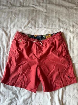 Rosa badshorts från Polo Ralph Lauren - Säljer ett par snygga rosa badshorts från Polo Ralph Lauren med elastisk midja och vit snörning. Shortsen har en bakficka med knapp och en liten Polo-logga framtill. Perfekta för sommaren och strandhäng!