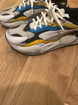 Färgglada sneakers från Puma - Säljer ett par chunky sneakers från Puma med vit, svart, gul och blå färg. Skorna har meshpartier, detaljer i syntet och skinn, samt vita snören. Snygg design med rund tå och platt sula, perfekt för dig som gillar streetstyle.