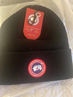 Svart mössa från Canada Goose - Ribbstickad svart mössa från Canada Goose med klassisk logotyp framtill. Perfekt för kalla dagar och håller dig varm med stil. Tidlös design som passar till det mesta.
