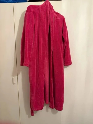 Rosa morgonrock  - Säljer en lång och mysig morgonrock i mjuk frotté. Västen har långa ärmar och ett matchande bälte i midjan för att enkelt kunna justera passformen. Perfekt för att chilla hemma eller efter duschen.
