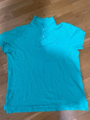 Turkos Polo Ralph Lauren t-shirt - Säljer en turkos t-shirt från Polo Ralph Lauren med klassisk pikékrage och två knappar framtill. T-shirten har korta ärmar och den ikoniska lilla broderade loggan på bröstet. Materialet är mjuk bomull och modellen är slim fit.