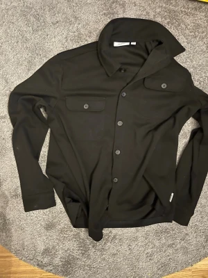 Svart overshirt från Only & Sons - Säljer en svart overshirt från Only & Sons med klassisk krage, två bröstfickor med lock och knappar framtill. Skjortan har lång ärm och är tillverkad i ett mjukt material som känns skönt mot huden. Perfekt att bära öppen över en t-shirt.